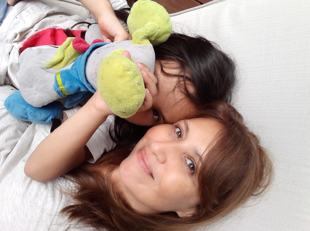Una mujer sonriente posando con una niña que sostiene un peluche en el sofá.