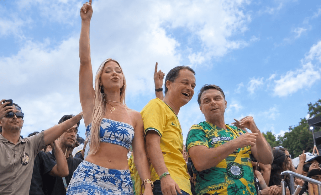 Grupo de personas celebrando en un evento al aire libre, con banderas y ropa de colores. Una mujer con top blanco y azul levanta un brazo, mientras dos hombres sonríen y lucen camisetas amarillas y verdes.