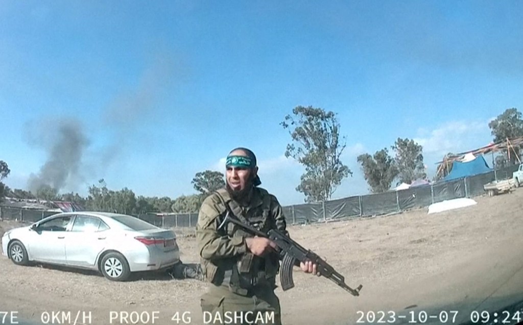 Un hombre vestido con uniforme militar sostiene un arma en un área al aire libre, con humo visible de un incendio en el fondo.